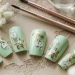 A set of mint green artificial nails featuring white flowers and gold leaf designs, arranged beside two nail art brushes and surrounded by delicate white flowers, perfect for showcasing elegant green nail art.
