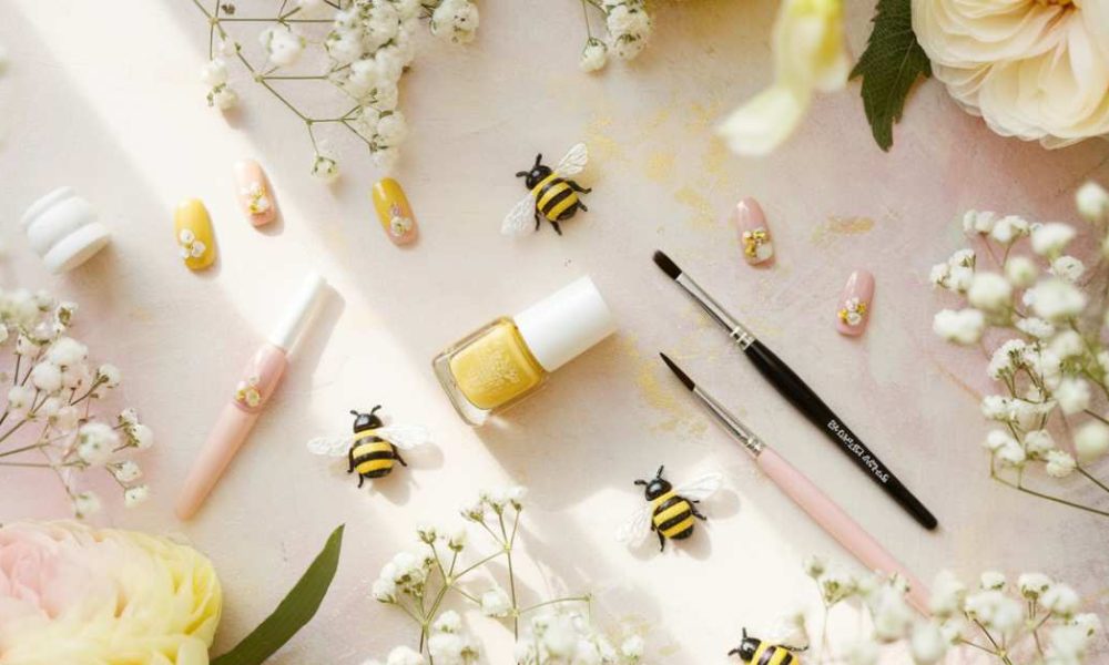 A flat lay of yellow nail polish, painted bee-themed fake nails, two brushes, small decorative bees, and white flowers on a soft, pastel-colored background. Surrounding are large cream and pink flowers.