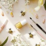 A flat lay of yellow nail polish, painted bee-themed fake nails, two brushes, small decorative bees, and white flowers on a soft, pastel-colored background. Surrounding are large cream and pink flowers.