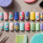 An array of colorful, artistic press-on nails with abstract designs showcases jelly nail art ideas, surrounded by various painting brushes and sponges in pastel hues on a textured surface.