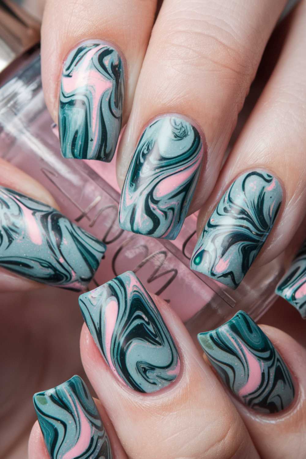 Close-up of a hand with long nails showcasing an intricate marbled design, a delightful inspiration for jelly nail art ideas. The pattern features swirling shades of teal, dark green, and pink on each nail. In the background is part of a nail polish bottle.