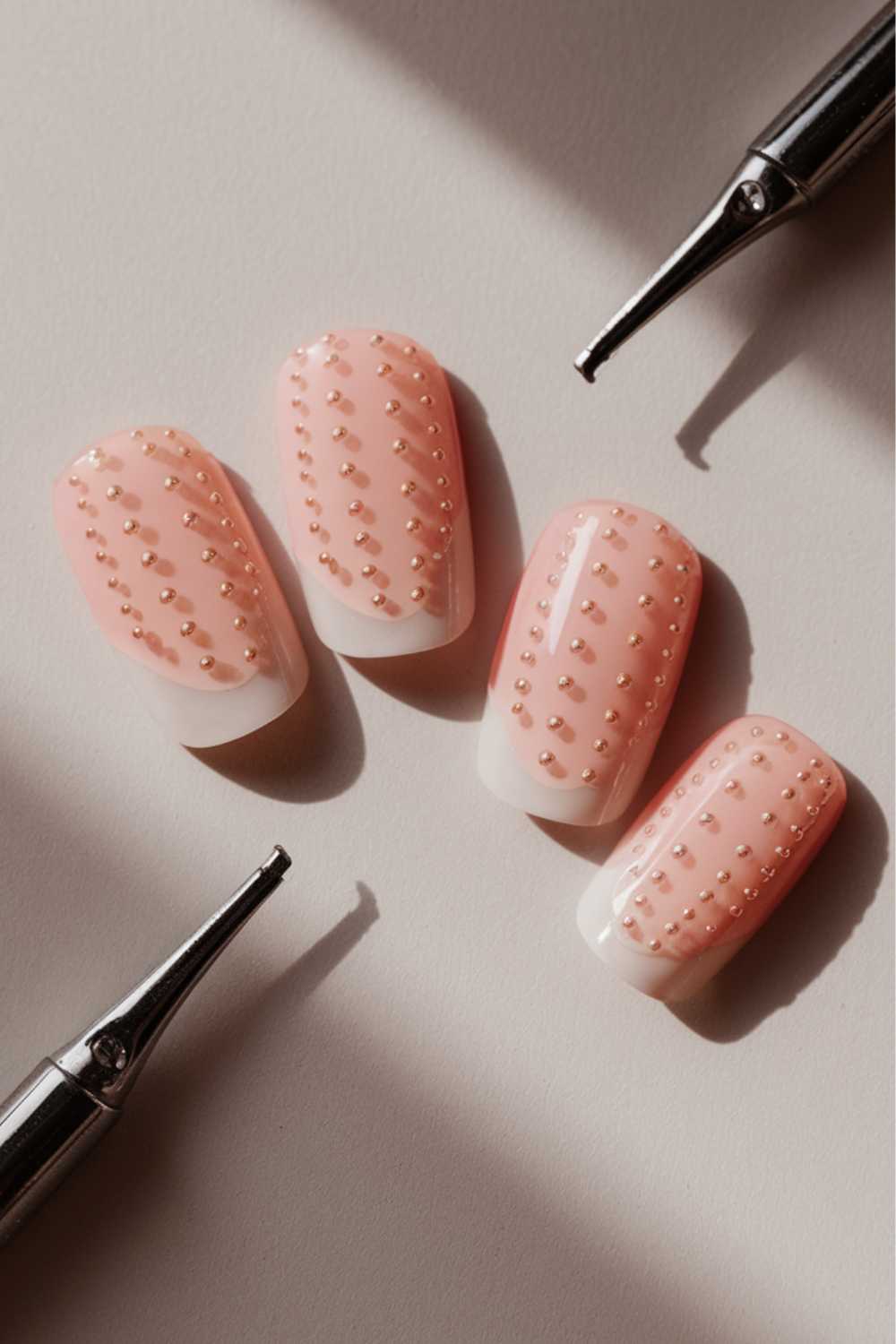 Four artificial nails are displayed on a light surface, painted in a jelly pink hue with small gold bead accents in neat rows. Two chrome nail tools rest nearby, casting small shadows, inspiring your next jelly nail art ideas.