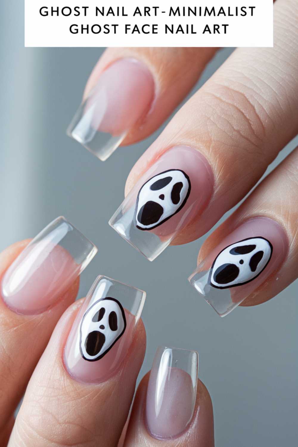 Close-up of a hand with clear, square-tipped nails featuring simple ghost face art on two nails. The design is minimalistic with black and white colors, giving a spooky effect. Caption at the top reads Ghost Nail Art - Minimalist Ghost Face Nail Art.