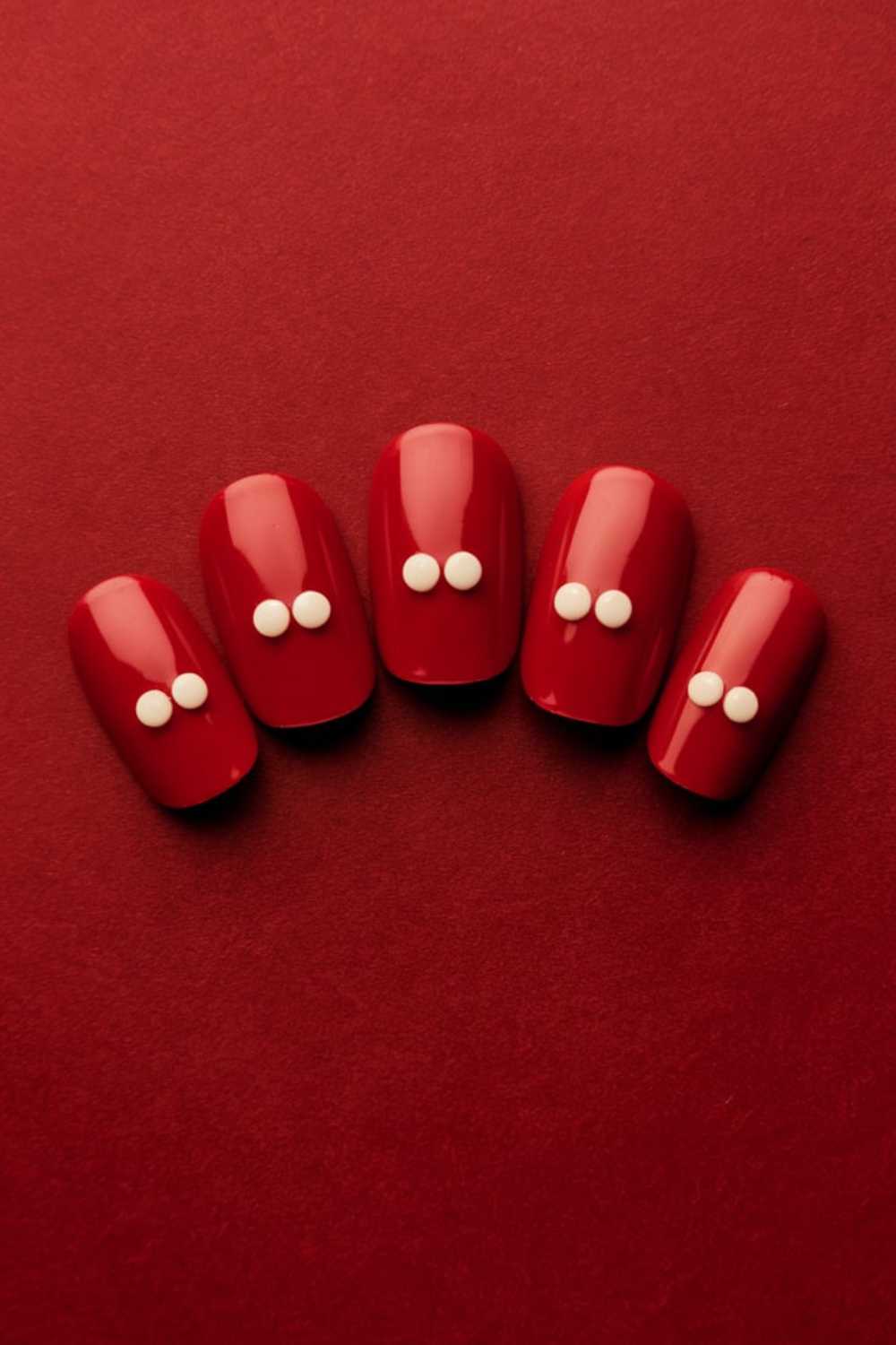 Red nails with a glossy finish are decorated with two small white dots on each nail, set against a matching red background.