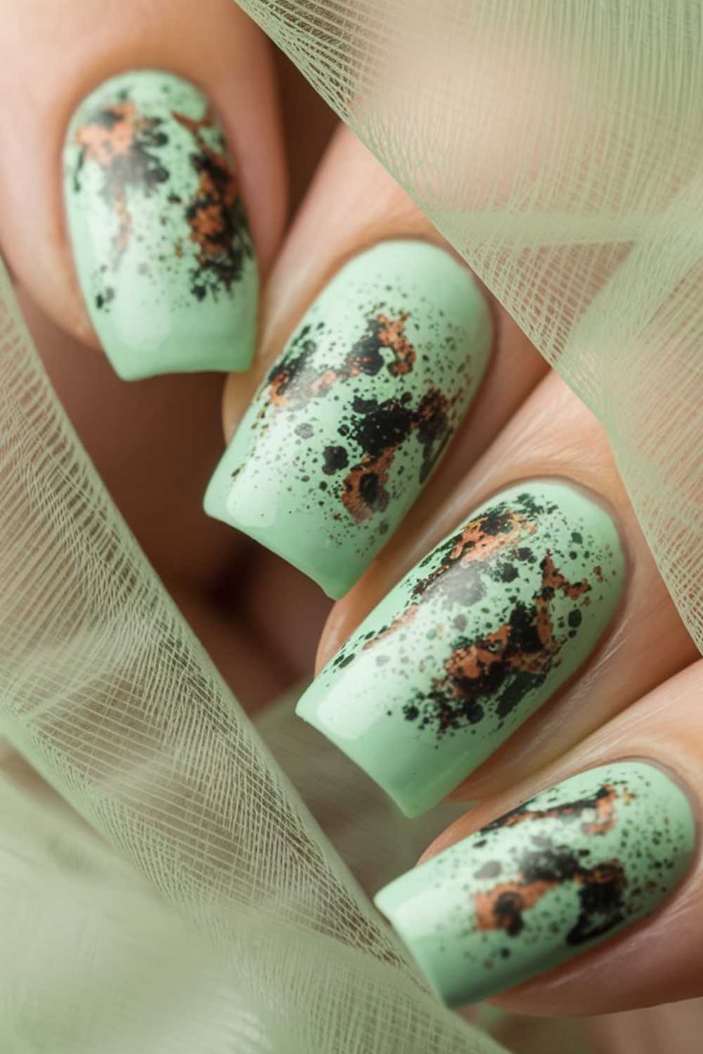 Close-up of fingernails painted mint green with black and copper splatter Green Nail Art, surrounded by soft, translucent green fabric.