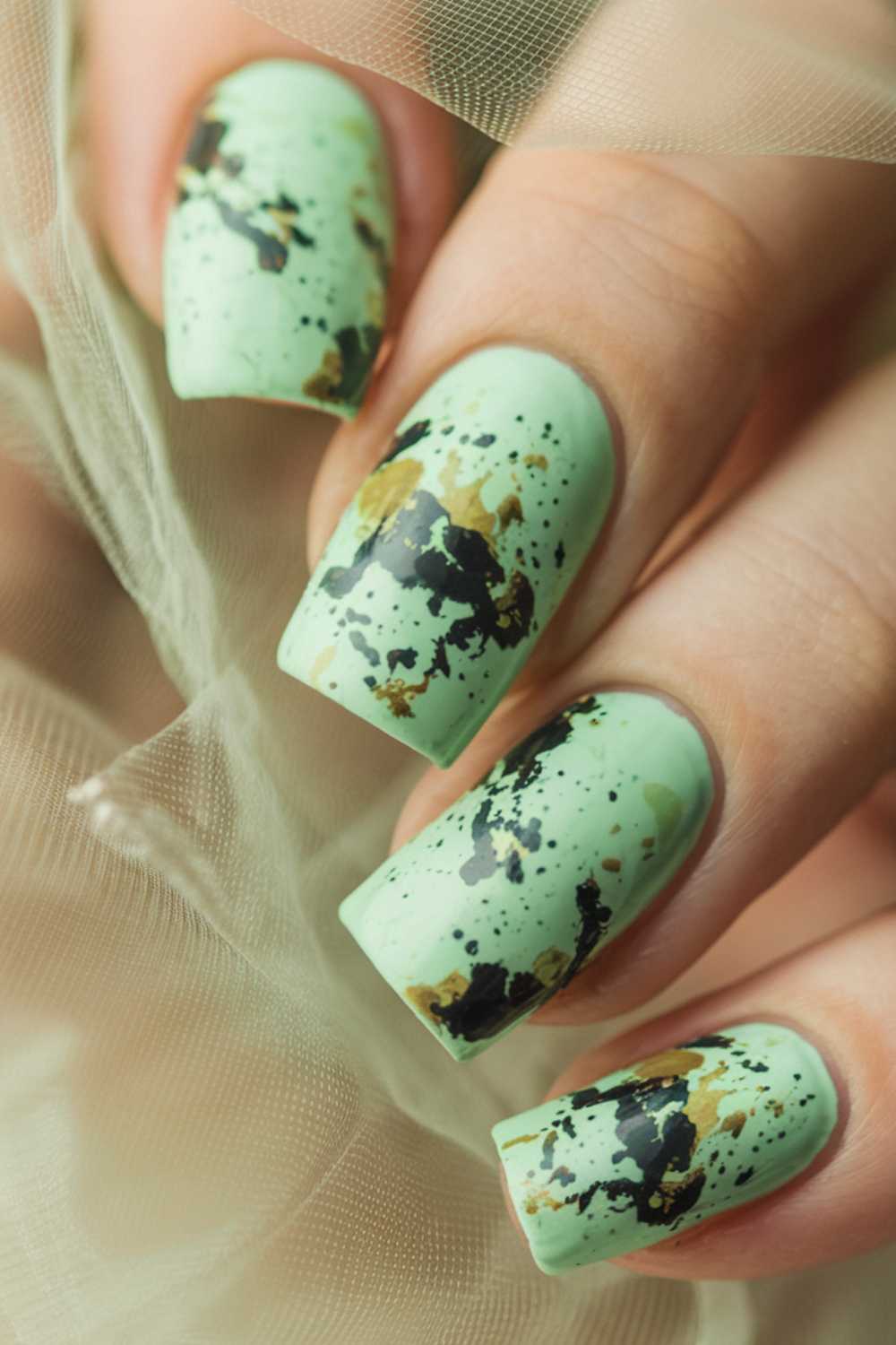 A close-up of a hand with long, square-shaped nails painted mint green, featuring bold green nail art and black and gold splatter accents. Sheer beige fabric surrounds the fingers.