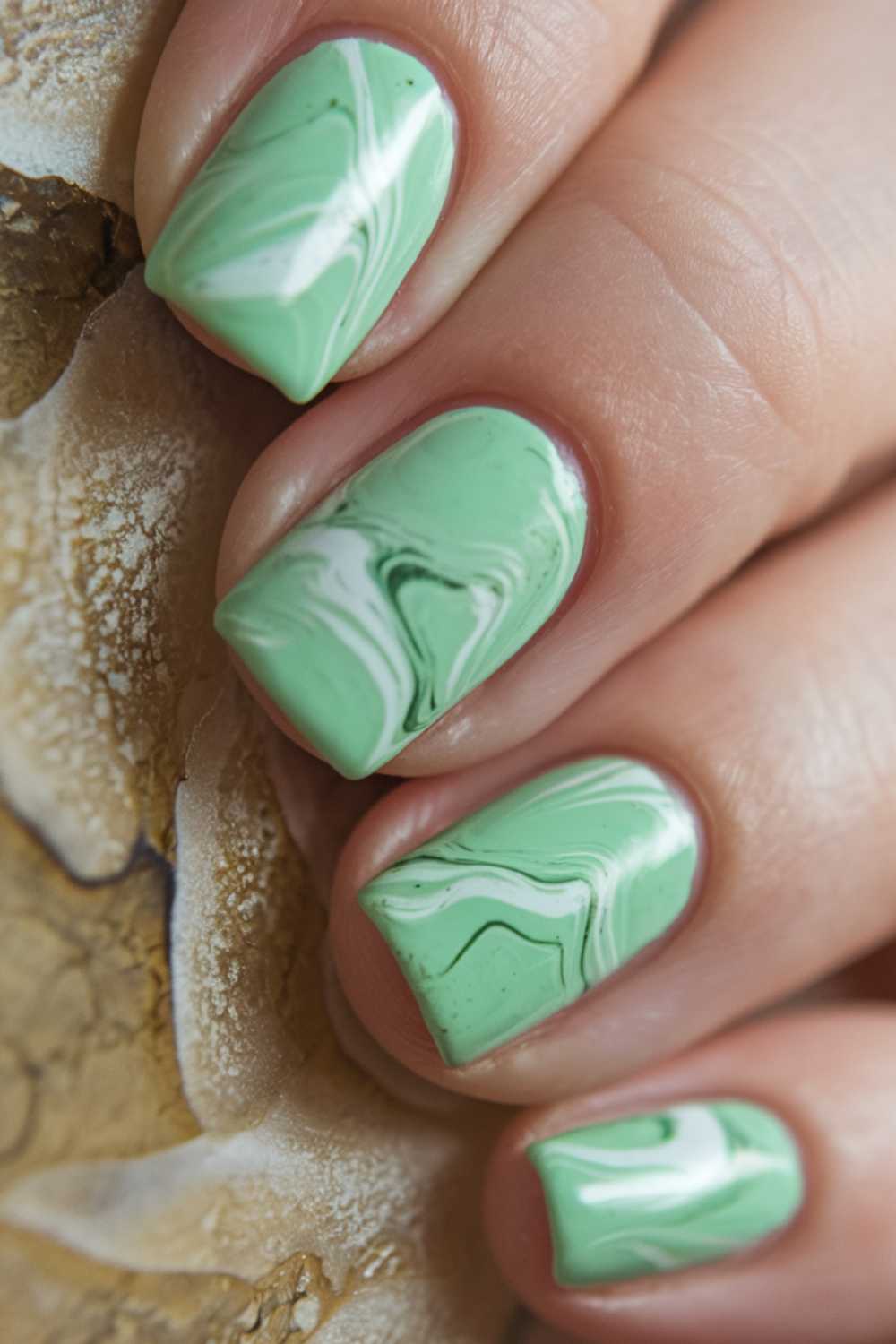 Close-up of fingernails painted with a light green marble effect, featuring swirling white and dark green patterns on each nail for an elegant Green Nail Art look.