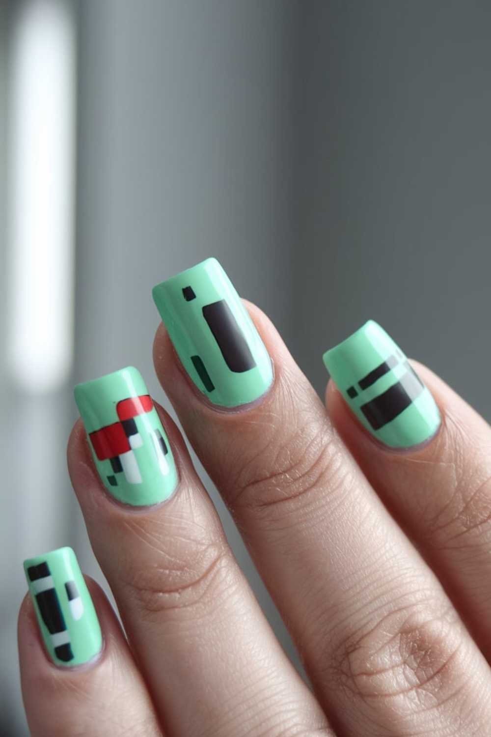 A hand with mint green nail polish showcases striking Green Nail Art, featuring geometric, abstract black, white, and red designs on each fingernail. The background is a softly blurred gray.
