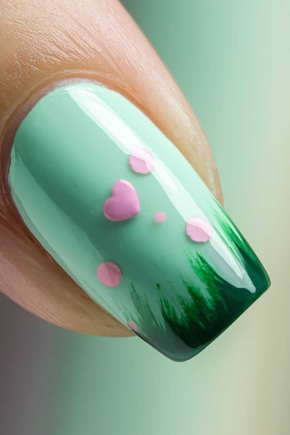 A close-up of Green Nail Art featuring a fingernail painted mint green with dark green at the tip, adorned with small pink dots and a pink heart.