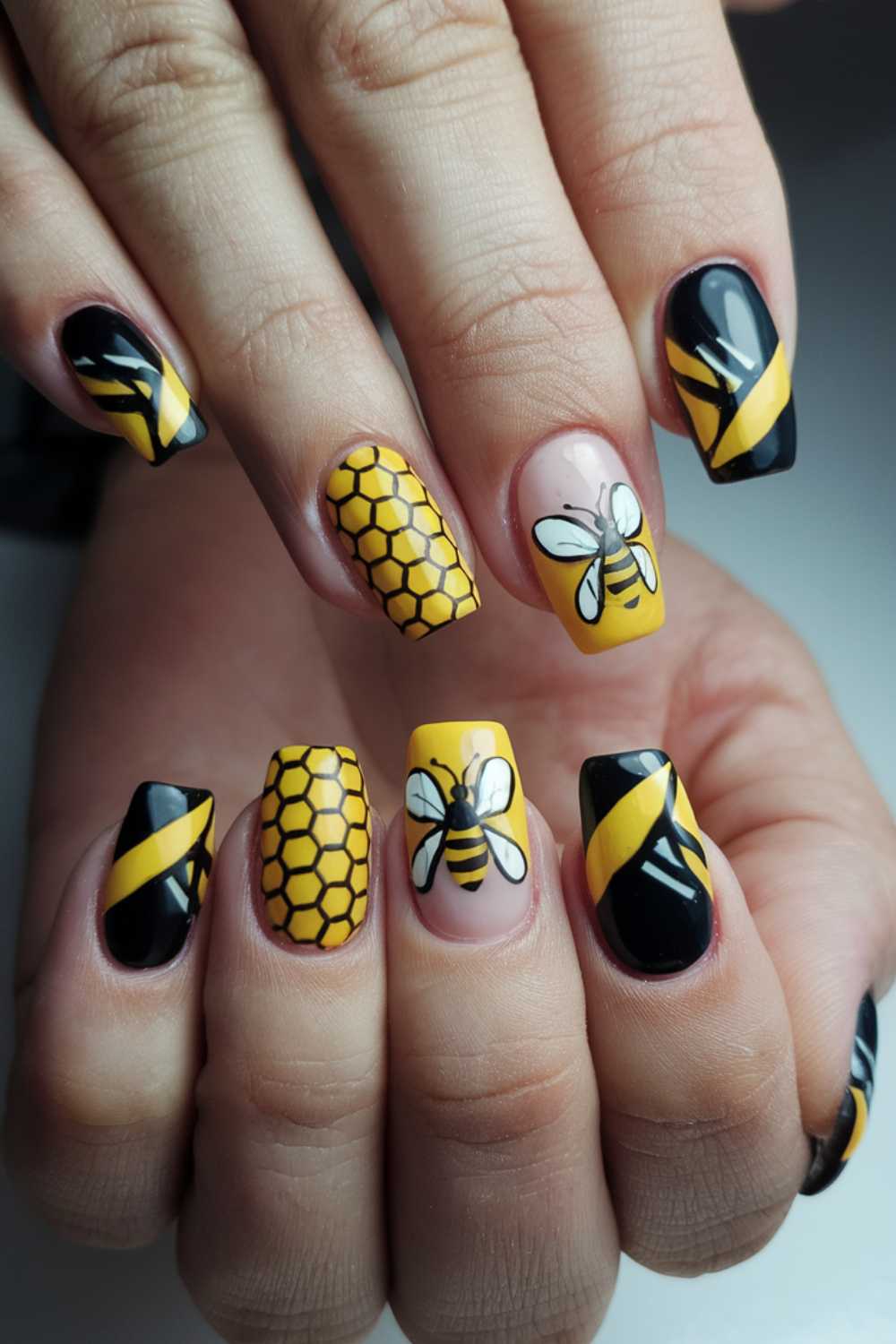 Close-up of hands showcasing exquisite bee-themed nail art. The nails feature intricate honeycomb patterns, black and yellow stripes, and detailed bee illustrations. Each Bee Nail Art design is distinct, blending black, yellow, and natural tones with artistic flair.