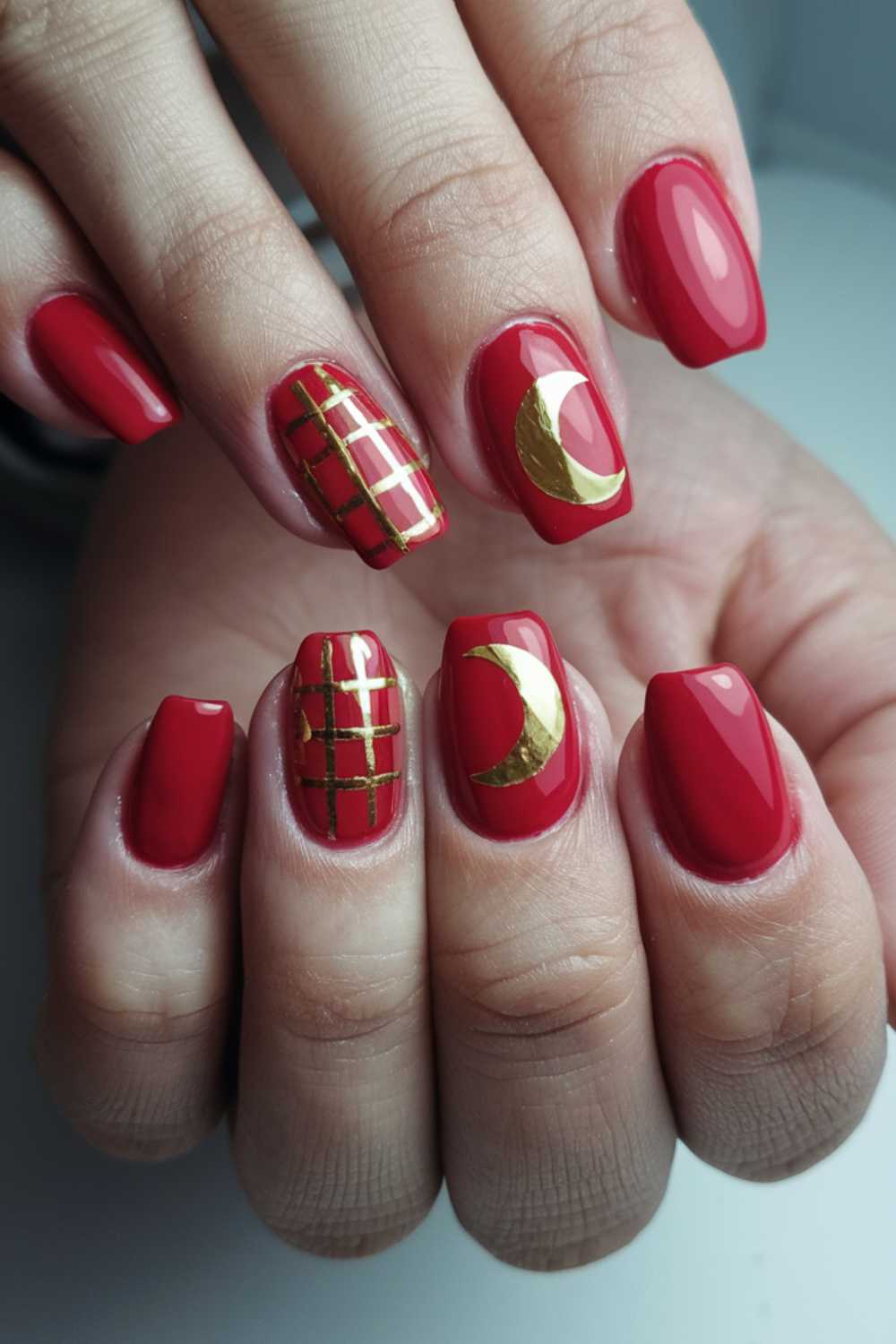 Close-up of hands with glossy red nails, featuring gold designs. The design includes a gold crescent and grid pattern on accent nails. The nails are shaped with a smooth, rounded finish.