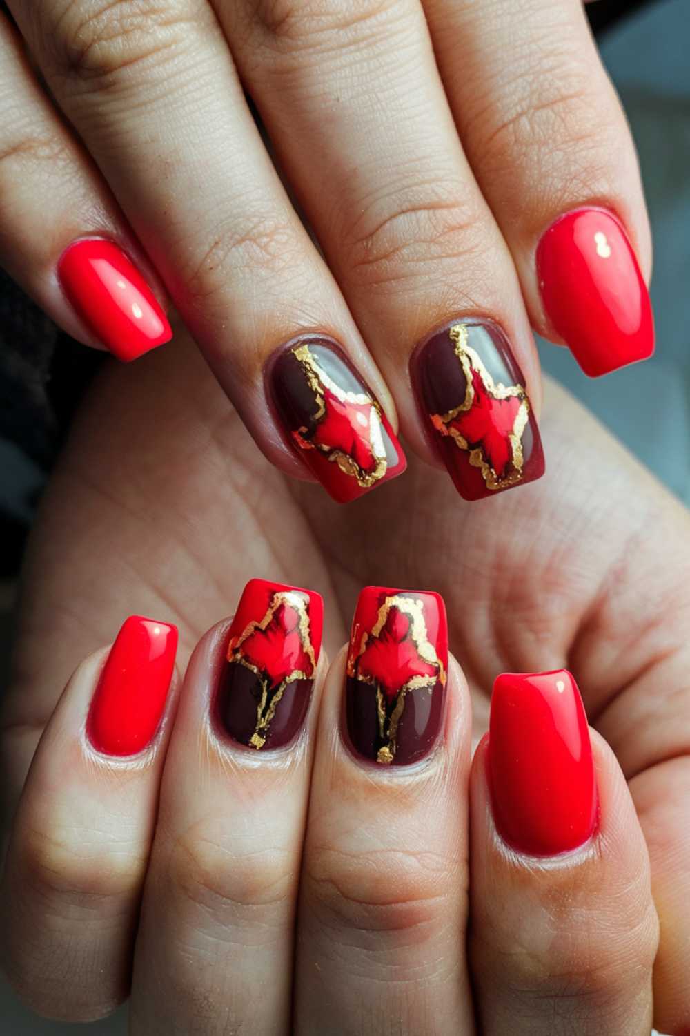 Close-up of freshly manicured hands with square-shaped nails. The nails are painted red, with accent nails featuring a dark brown base, red star patterns, and gold details, creating a bold and artistic design.