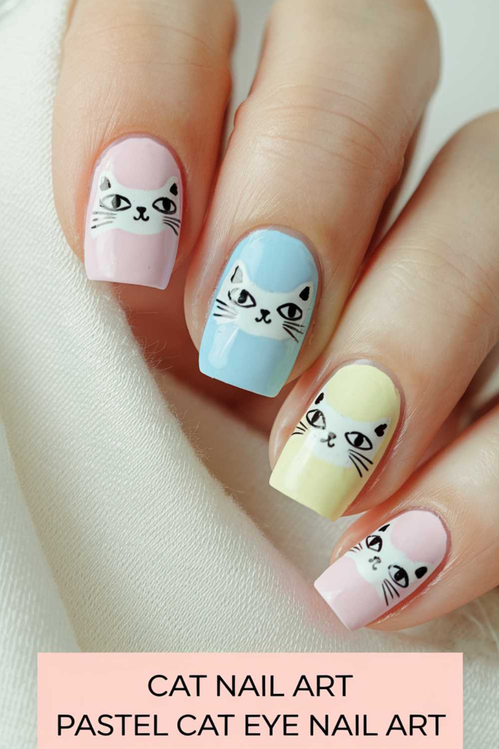 Close-up of a hand with pastel cat-eye nail art. Each nail is painted a different pastel color—pink, blue, yellow, and pink—with a cartoon cat face design. A white fabric is draped in the background. Text at the bottom reads CAT NAIL ART PASTEL CAT EYE NAIL ART.