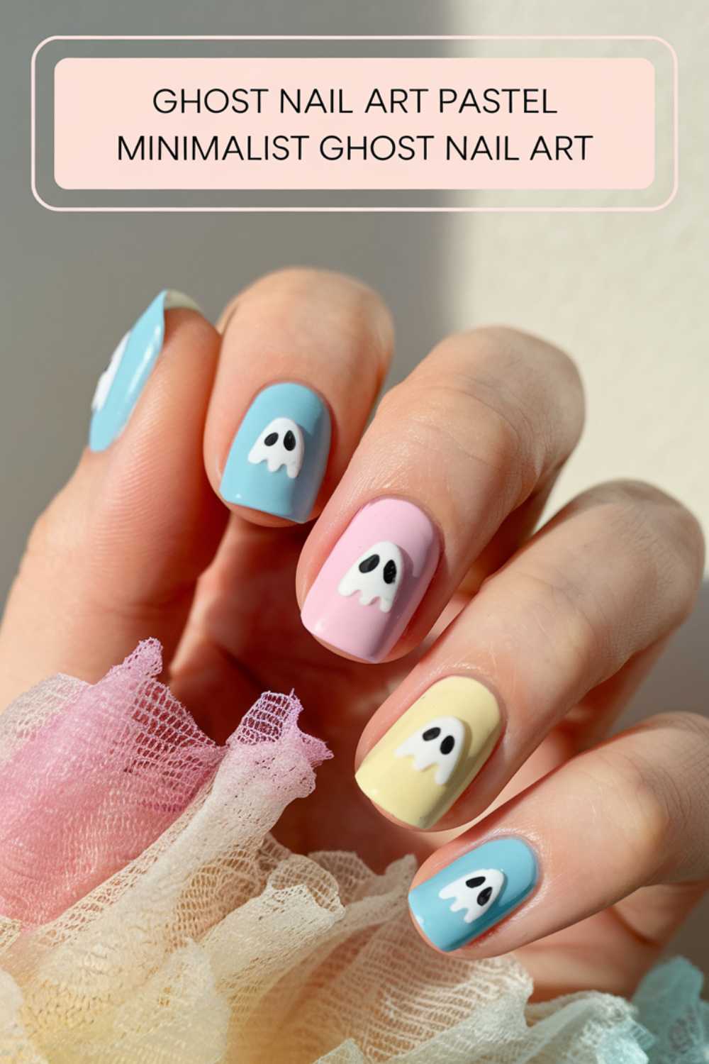 Close-up of a hand with pastel nail art featuring minimalist ghost designs on blue, pink, and yellow backgrounds. The nails have cute ghost faces, and the hand is resting on soft, multicolored tulle.