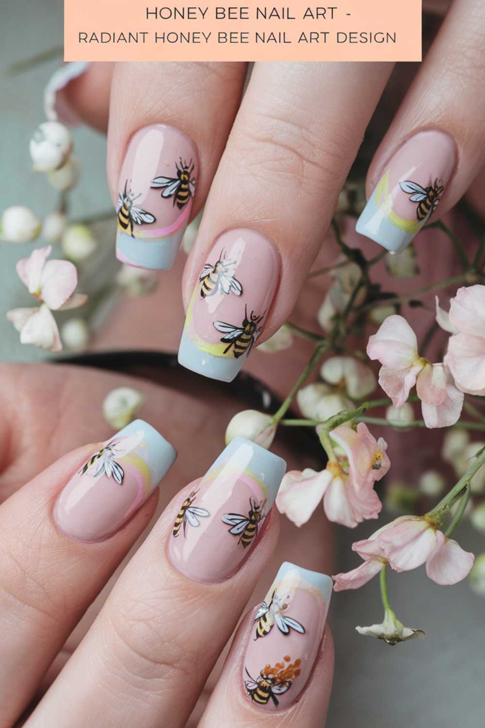 Close-up of a hand with bee nail art. Nails gleam with blue tips, showcasing intricate designs of bees fluttering near painted flowers. Delicate pink blooms are held alongside, enhancing the detailed artistry. Text at the top reads: Honey Bee Nail Art - Radiant Bee Nail Art Design.