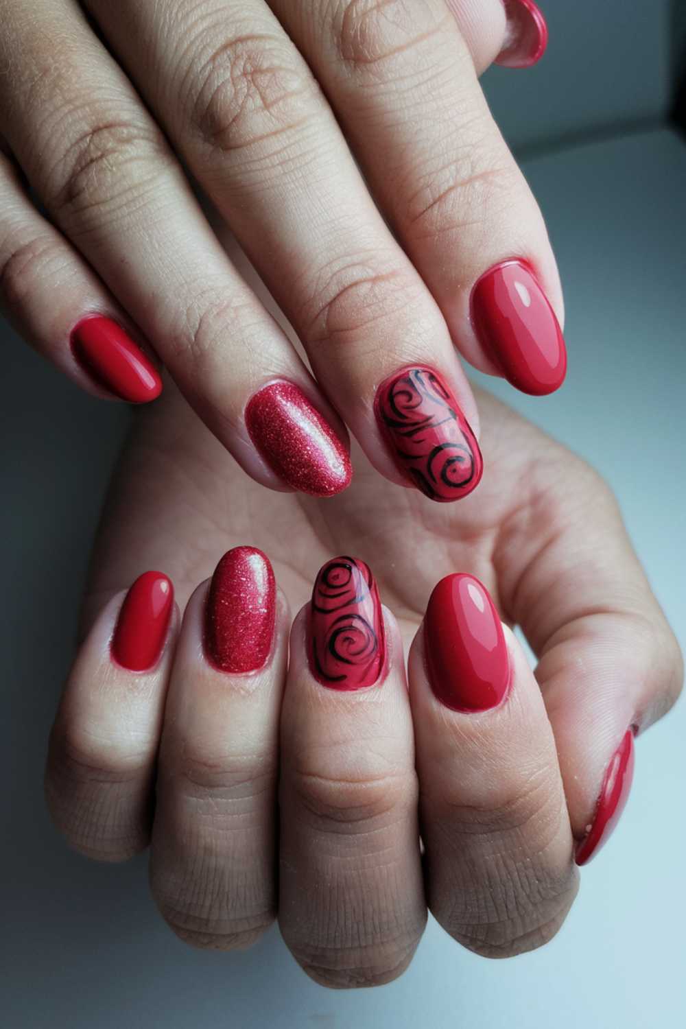 Close-up of hands with red manicured nails. Most nails are shiny red; one has red glitter, and two feature black floral patterns on a red base. The fingers are arranged artistically, showcasing the nail designs.