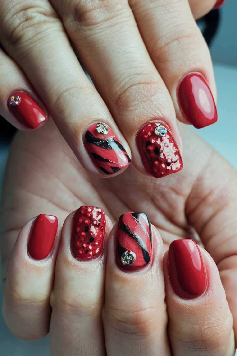 Close-up of hands with red manicured nails featuring glossy polish. Some nails have black stripes and others have small studs for added texture. The design includes embellishments like tiny gems for a decorative effect.