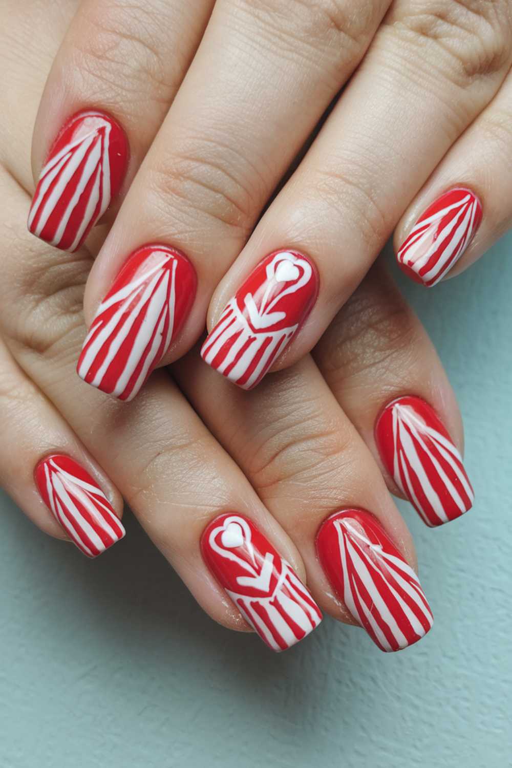Close-up of hands with festive nail art. Nails are painted red with white striped designs resembling candy canes. The ring fingers feature a heart on top of the striped pattern, against a light teal background.