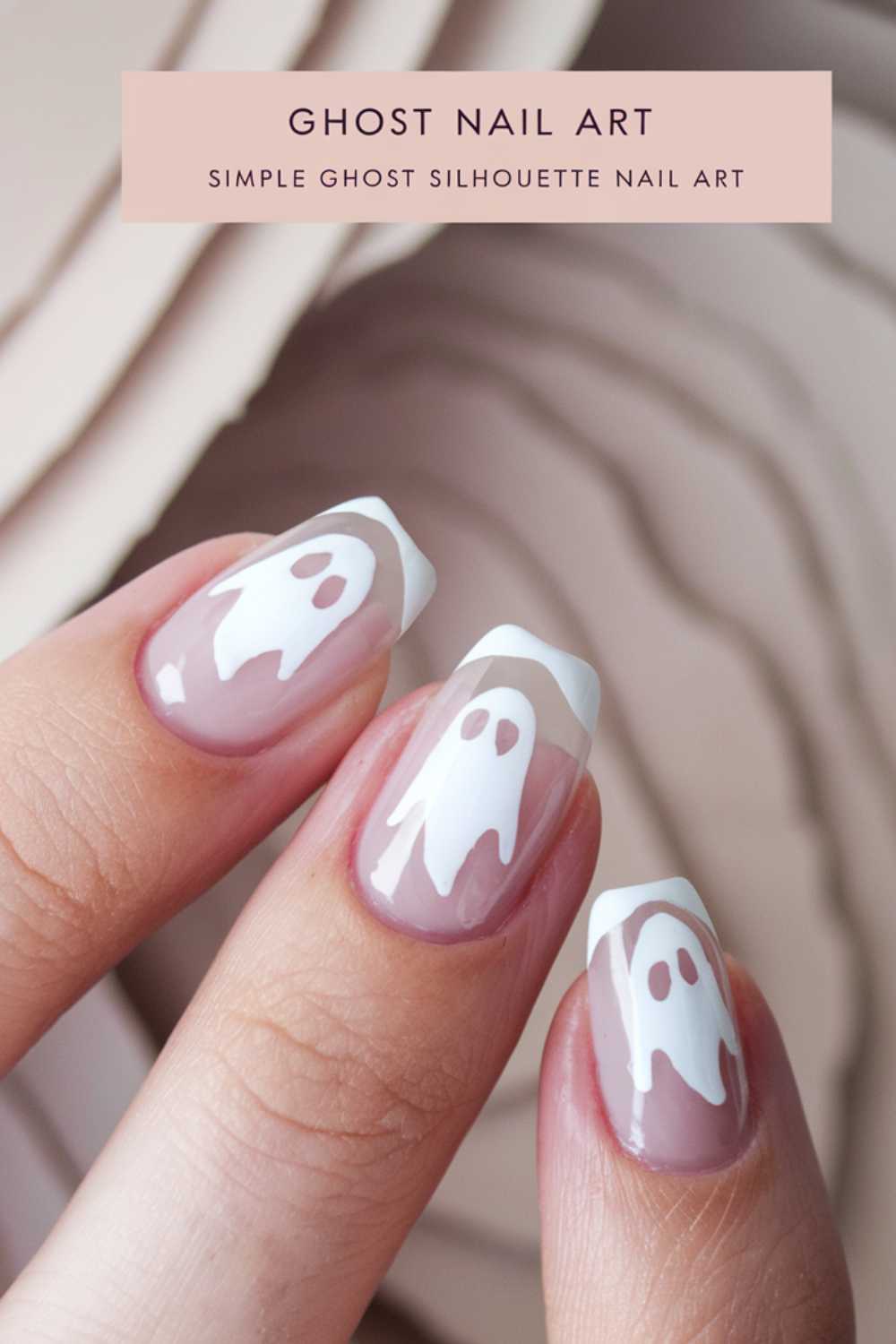 Close-up of a hand with ghost-themed nail art. Each fingernail shows a white ghost silhouette on a natural pink base with a white tip. Background features a blurred neutral-toned pattern.