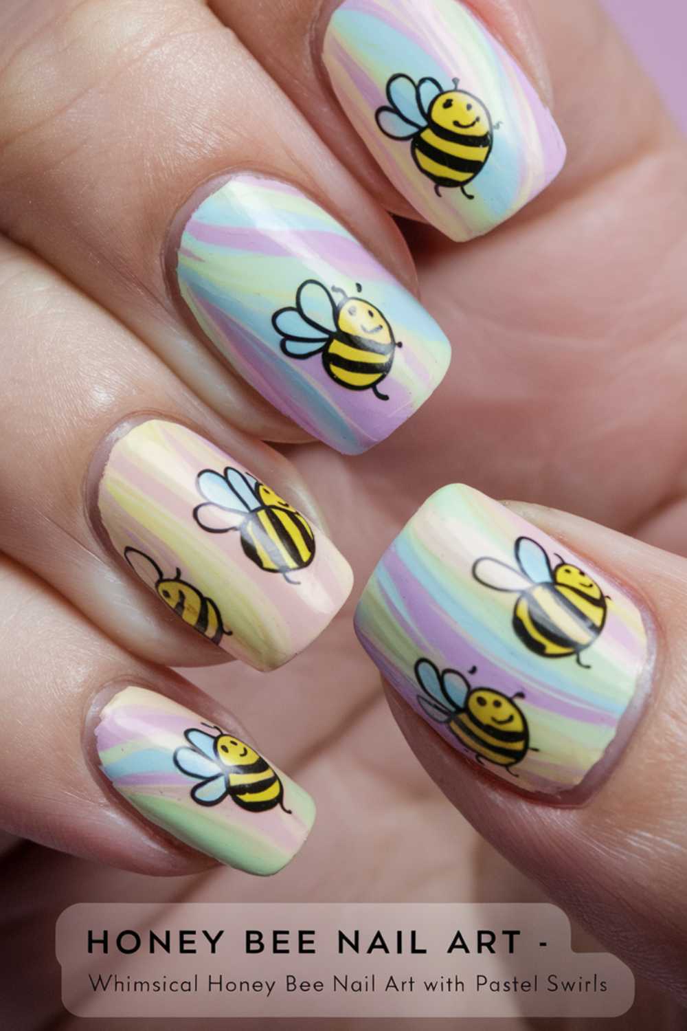 Close-up of fingers showcasing bee nail art, featuring cheerful honey bees in cartoon style with yellow and black stripes. The pastel swirl background blends soft pink, blue, green, and yellow hues for a playful touch.