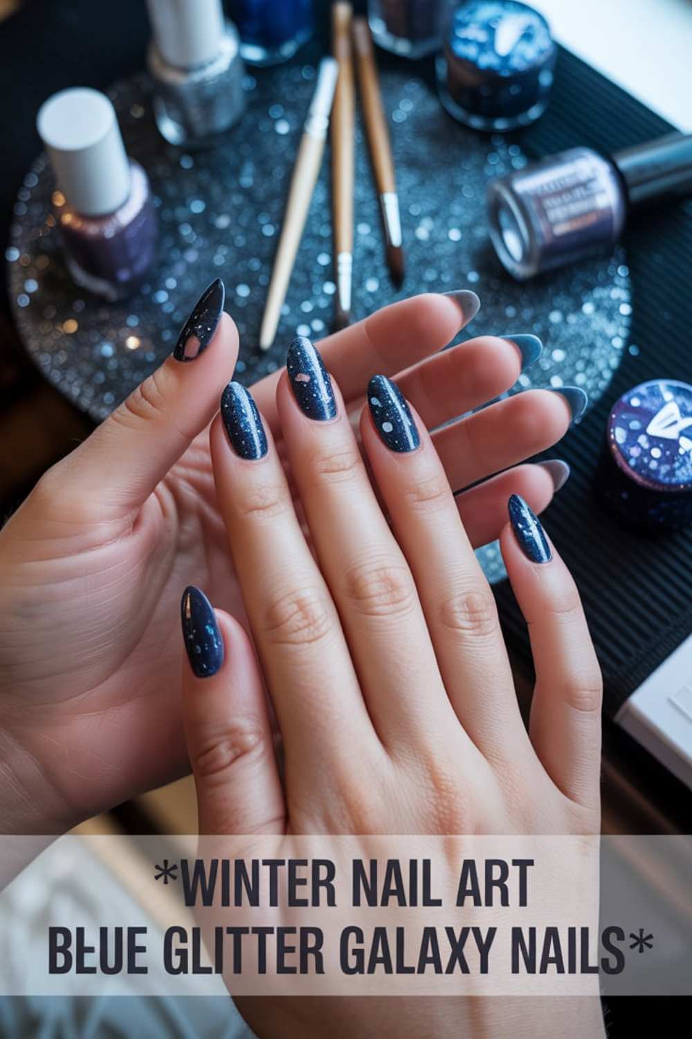 A person displays long, almond-shaped nails painted dark blue with white speckles, resembling a starry galaxy. Nail polish bottles and brushes are nearby. Text reads: Winter Nail Art Blue Glitter Galaxy Nails.