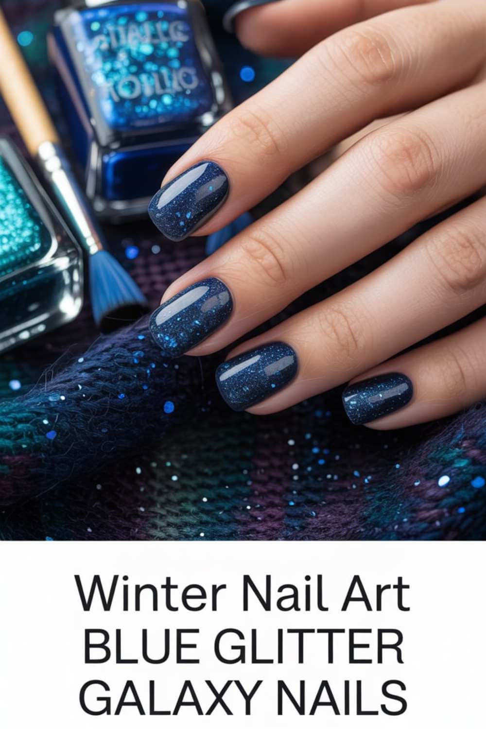 A hand with short, square-shaped nails painted in dark blue glitter polish. Bottles of blue nail polish and a brush are nearby. The text reads, Winter Nail Art BLUE GLITTER GALAXY NAILS.