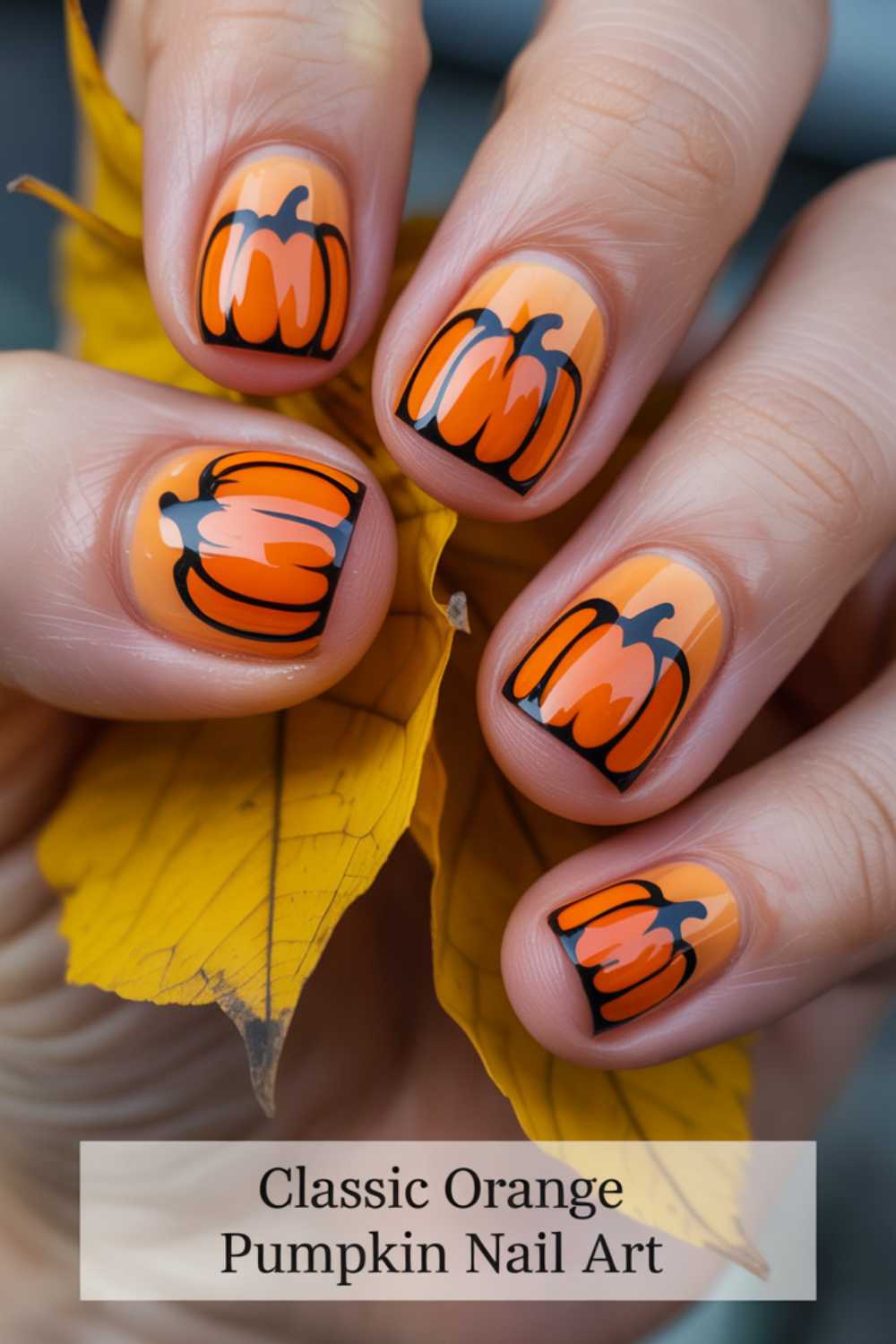 A hand with short, rounded nails painted with orange pumpkin nail art rests on a yellow autumn leaf. Text at the bottom reads: “Classic Orange Pumpkin Nail Art.”.