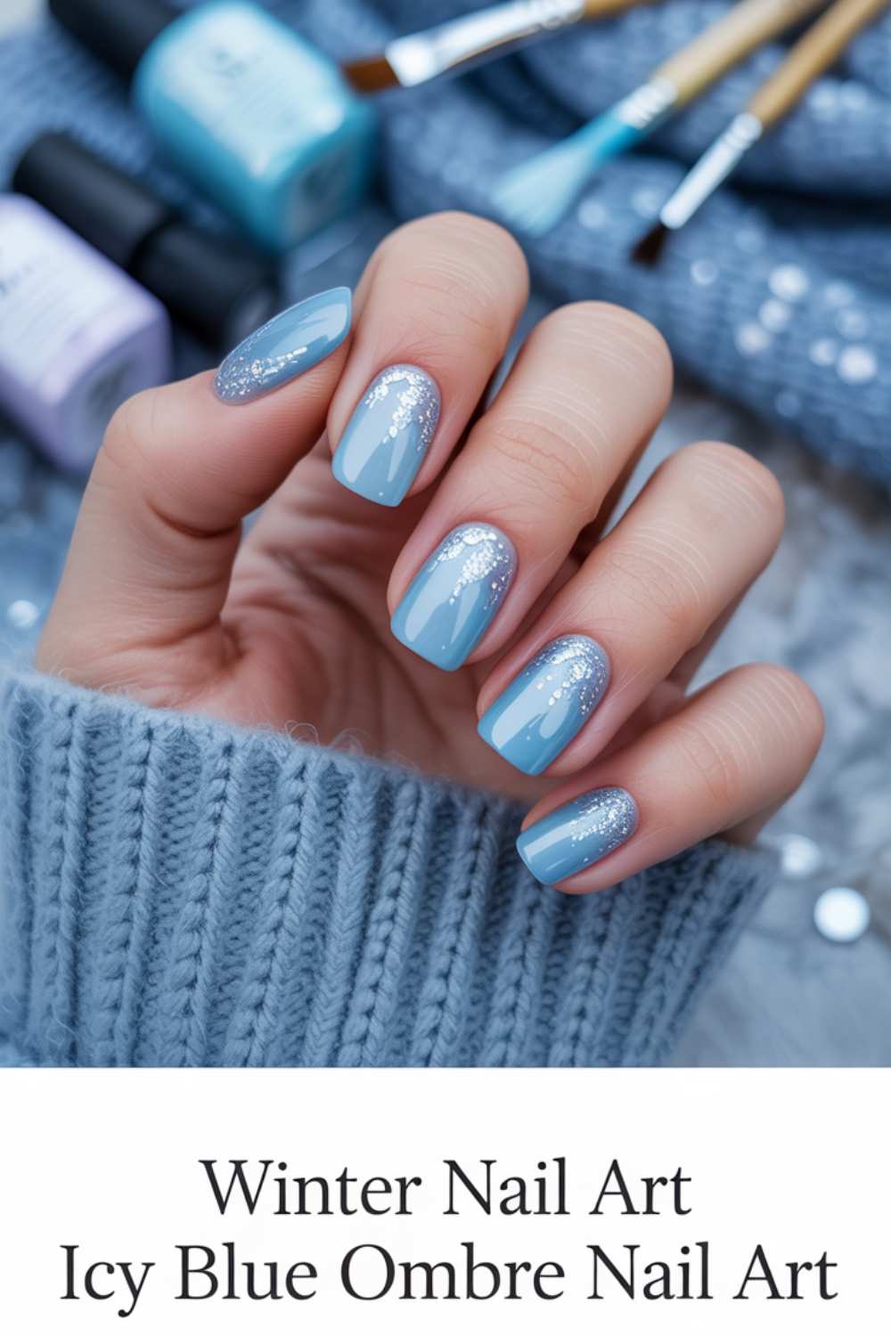 A hand with icy blue ombre nail art and sparkly silver glitter accents, wearing a cozy blue sweater. Nail polish bottles and brushes are in the blurred background. Text reads: Winter Nail Art Icy Blue Ombre Nail Art.