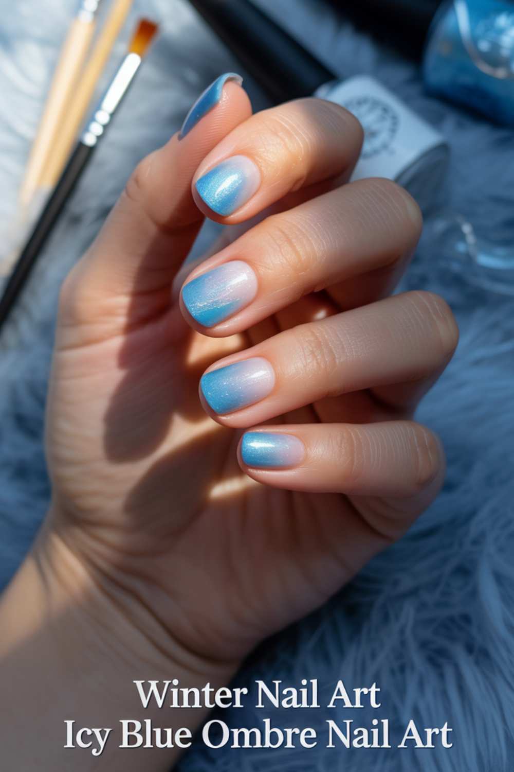 A hand with short, oval nails features an icy blue ombre nail art design. The background is soft gray faux fur, and text at the bottom reads: “Winter Nail Art Icy Blue Ombre Nail Art.” Paintbrushes are visible in the background.
