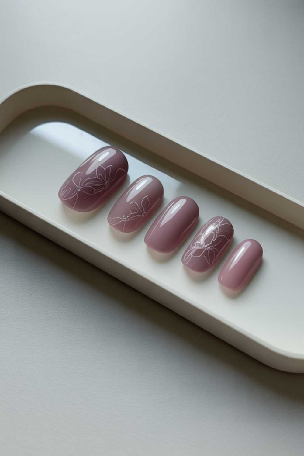 A set of five artificial mauve-colored nails with a glossy finish, displayed on a cream tray. Some nails feature delicate white floral line art designs.