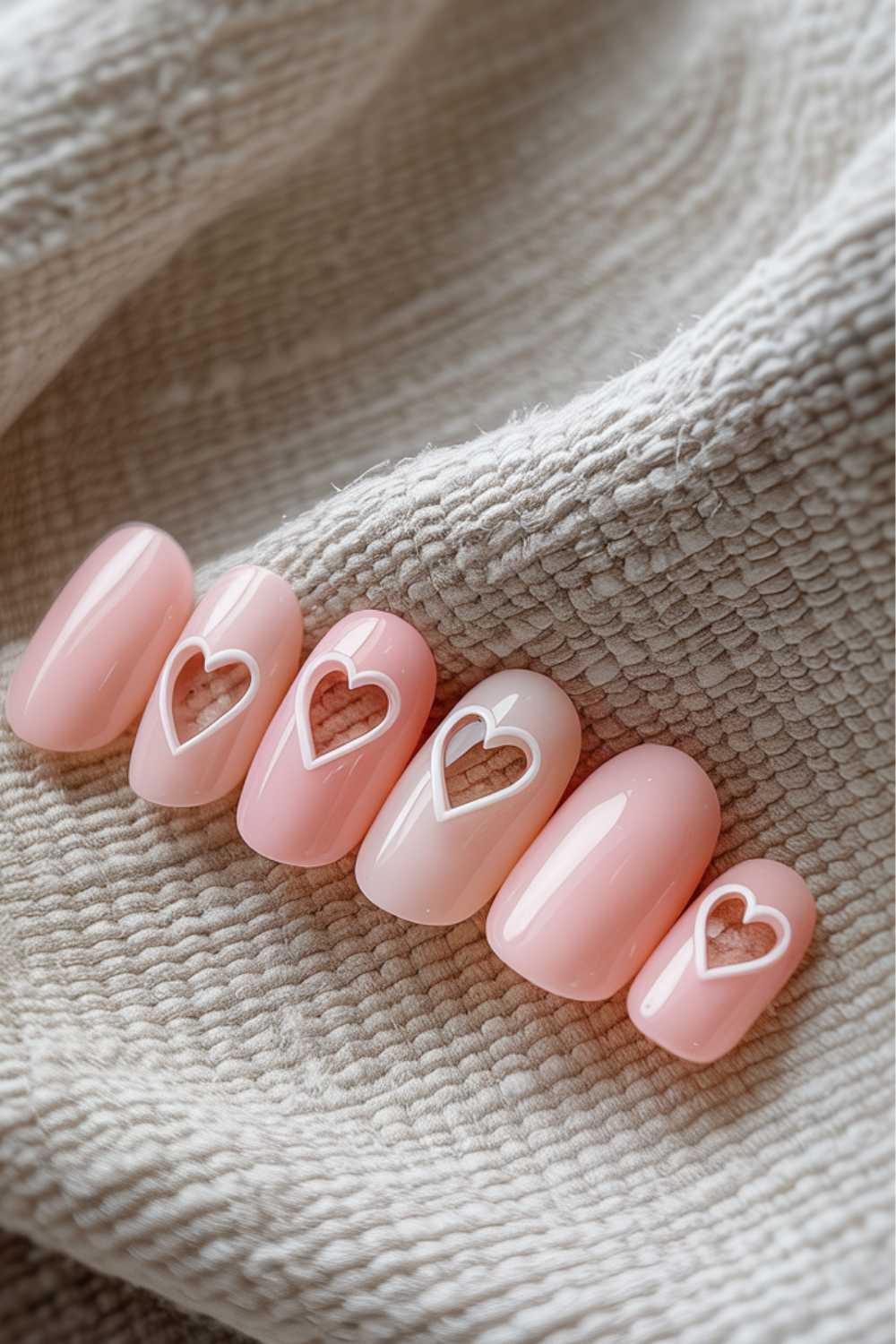 Five artificial nails with a pink, glossy finish are displayed, each featuring a white heart outline design. The nails are arranged on a textured beige fabric background.