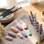 A set of pastel-colored false nails is arranged on a fabric napkin beside dried flowers, paintbrushes, and small plates on a wooden table in soft natural light.