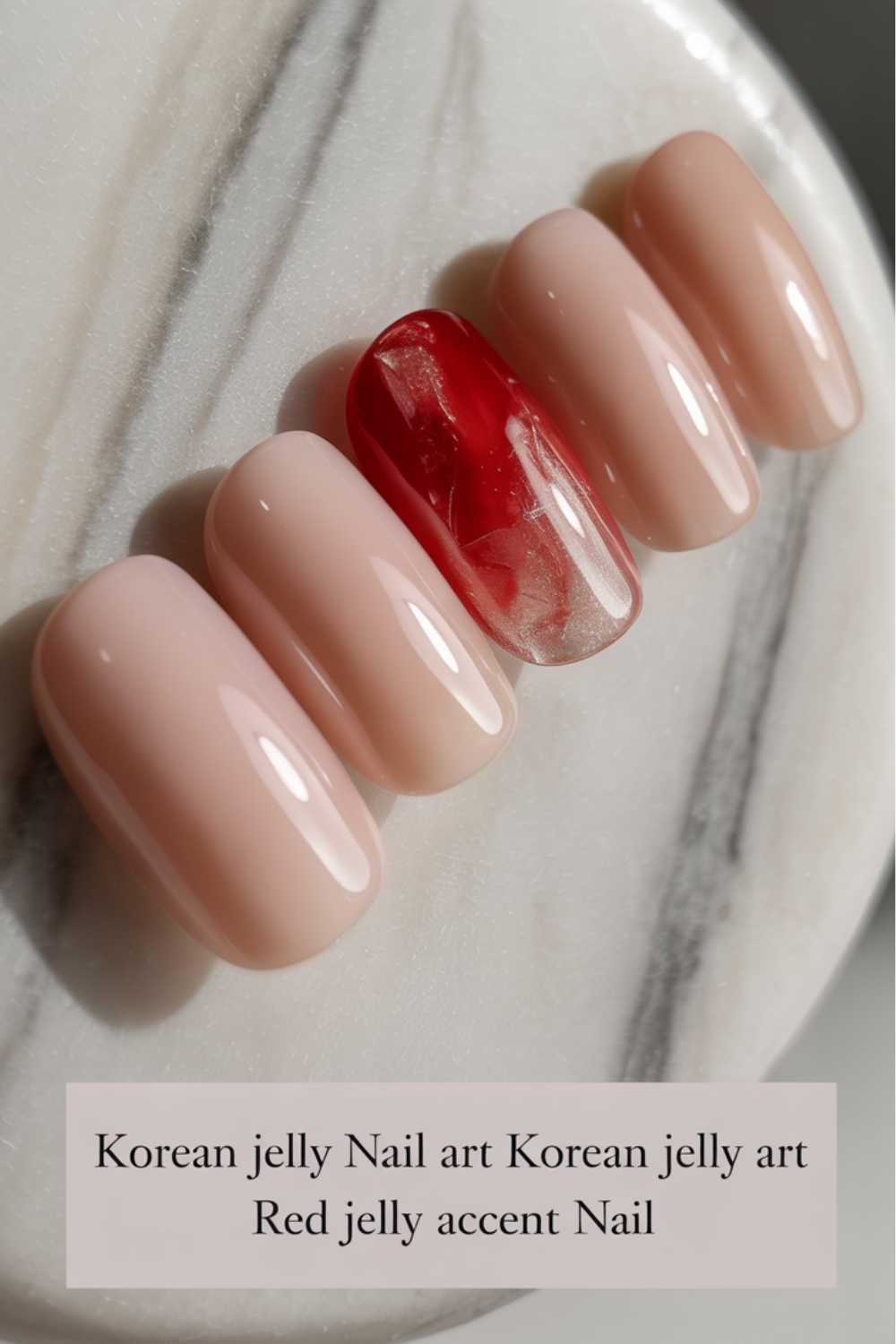 Five artificial nails displayed on a marble surface, four painted in a light nude color and one in the center featuring a red jelly accent with a translucent, marbled effect.