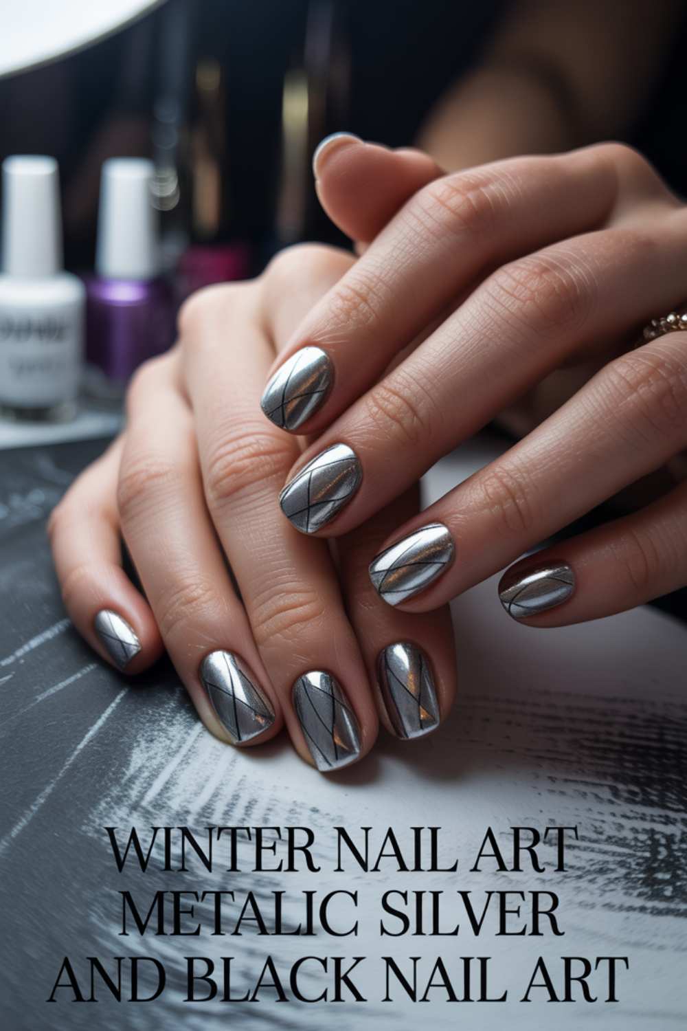 Close-up of hands with metallic silver and black geometric nail art. Nails are shiny with crisscrossed black lines, resting on a stylish background. Bottles of nail polish are visible in the background. Text reads Winter Nail Art Metallic Silver and Black Nail Art.