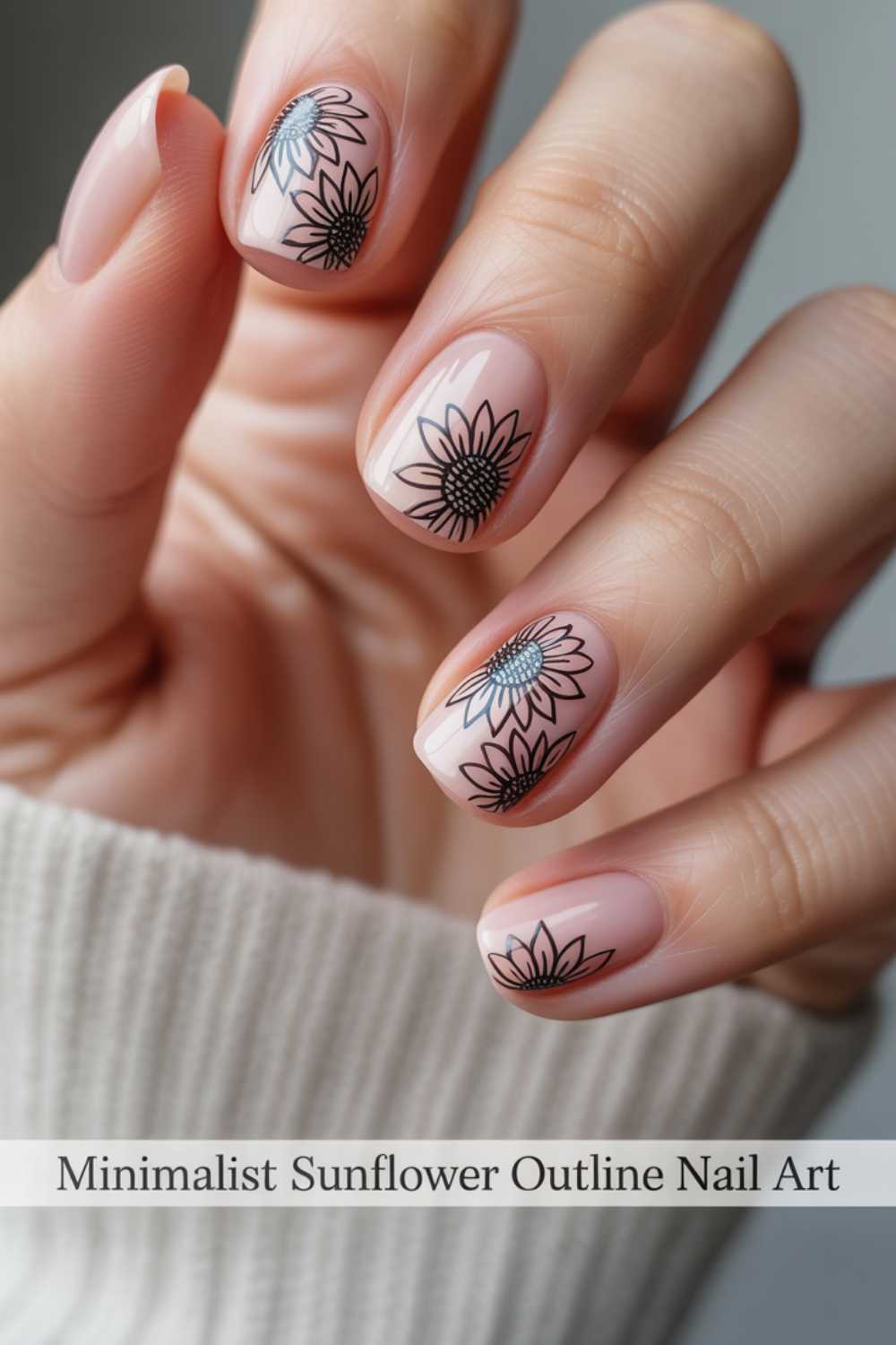 A hand with nude-colored nails featuring minimalist black sunflower outline designs on each nail. The person is wearing a light-colored sweater. Text at the bottom reads, Minimalist Sunflower Outline Nail Art.