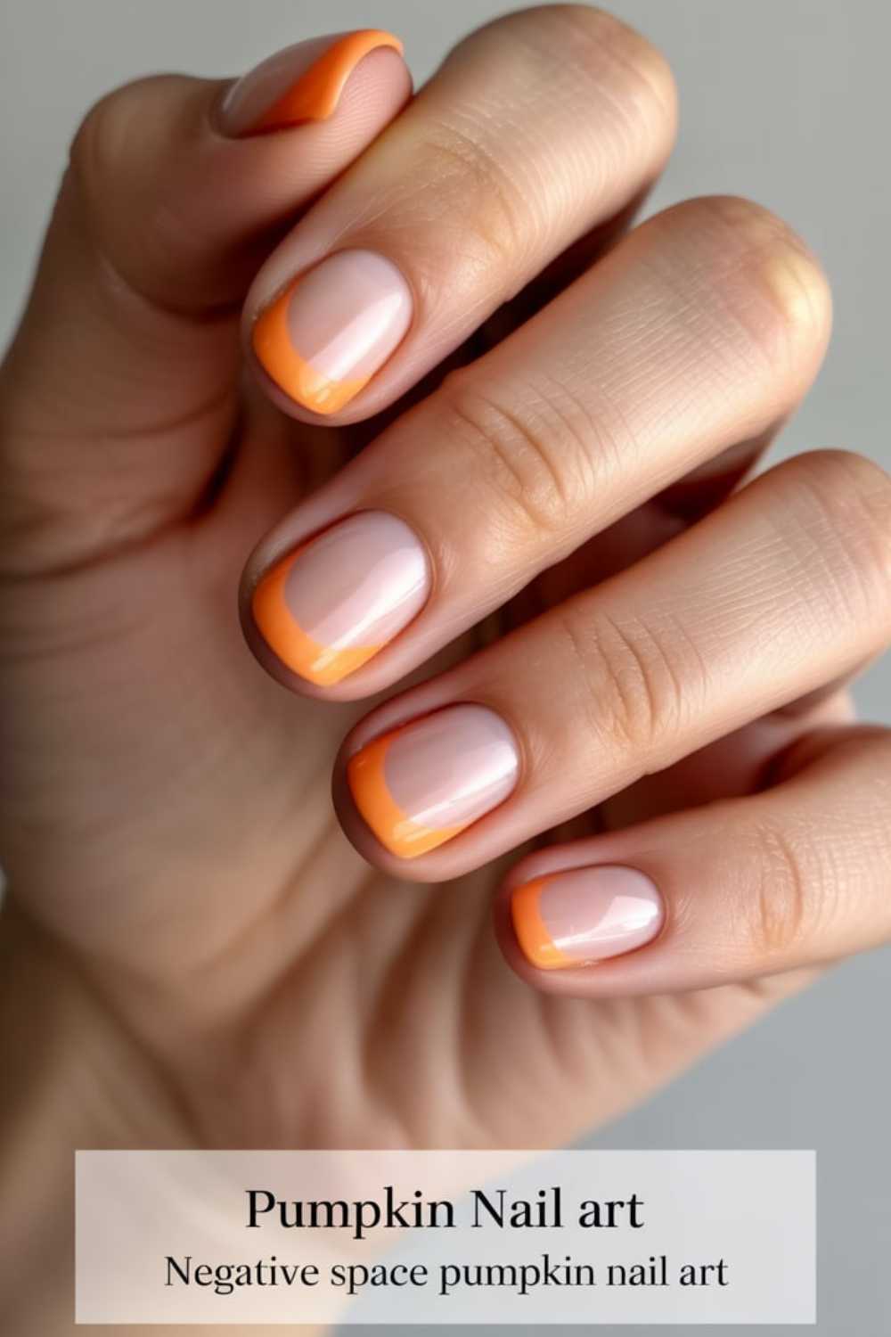 A hand with neatly manicured nails featuring negative space nail art. Each nail has an orange French tip with an extra curved pumpkin-orange line, resembling a pumpkin-inspired design. Text: Pumpkin Nail art. Negative space pumpkin nail art.