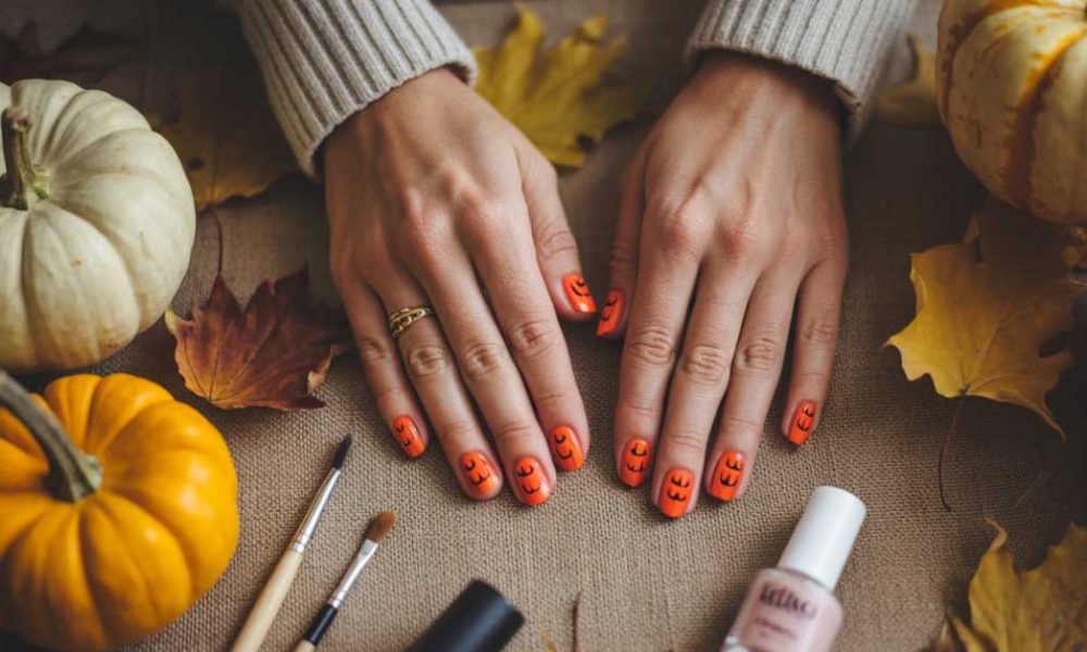 A pair of hands with orange Halloween-themed nail art featuring jack-o’-lantern faces rests on a table surrounded by small pumpkins, autumn leaves, makeup brushes, and nail polish.