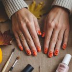 A pair of hands with orange Halloween-themed nail art featuring jack-o’-lantern faces rests on a table surrounded by small pumpkins, autumn leaves, makeup brushes, and nail polish.