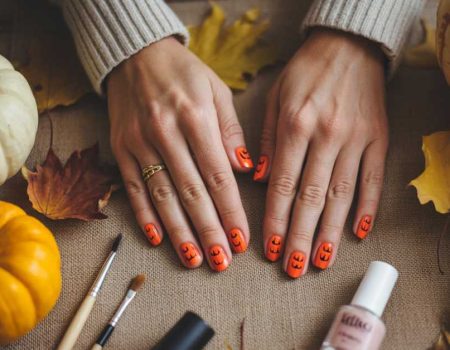 A pair of hands with orange Halloween-themed nail art featuring jack-o’-lantern faces rests on a table surrounded by small pumpkins, autumn leaves, makeup brushes, and nail polish.