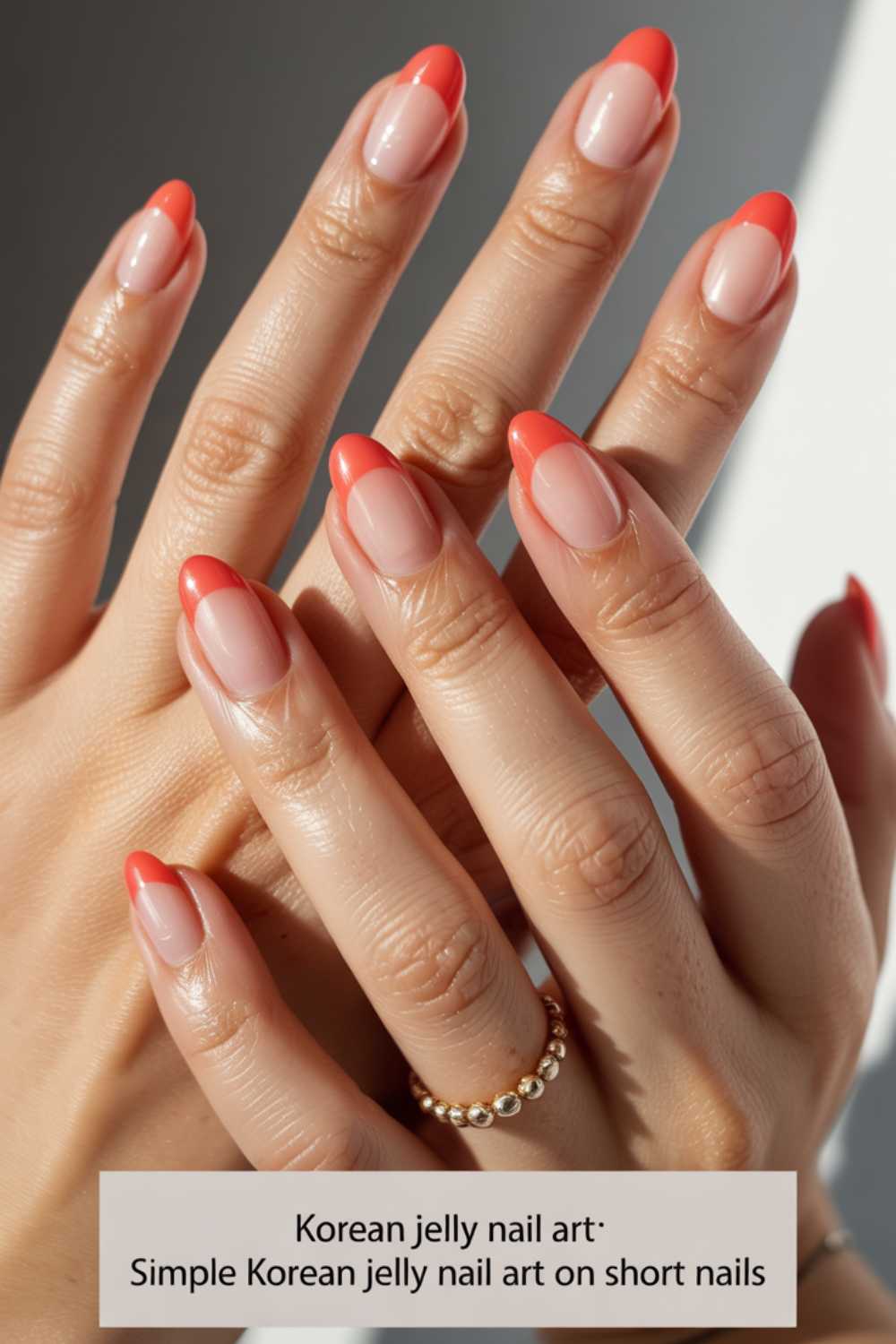 Close-up of hands with short nails featuring Korean jelly nail art, showcasing a translucent nude base and bright coral tips. A gold beaded ring is worn on one finger. Text at the bottom describes the nail style.