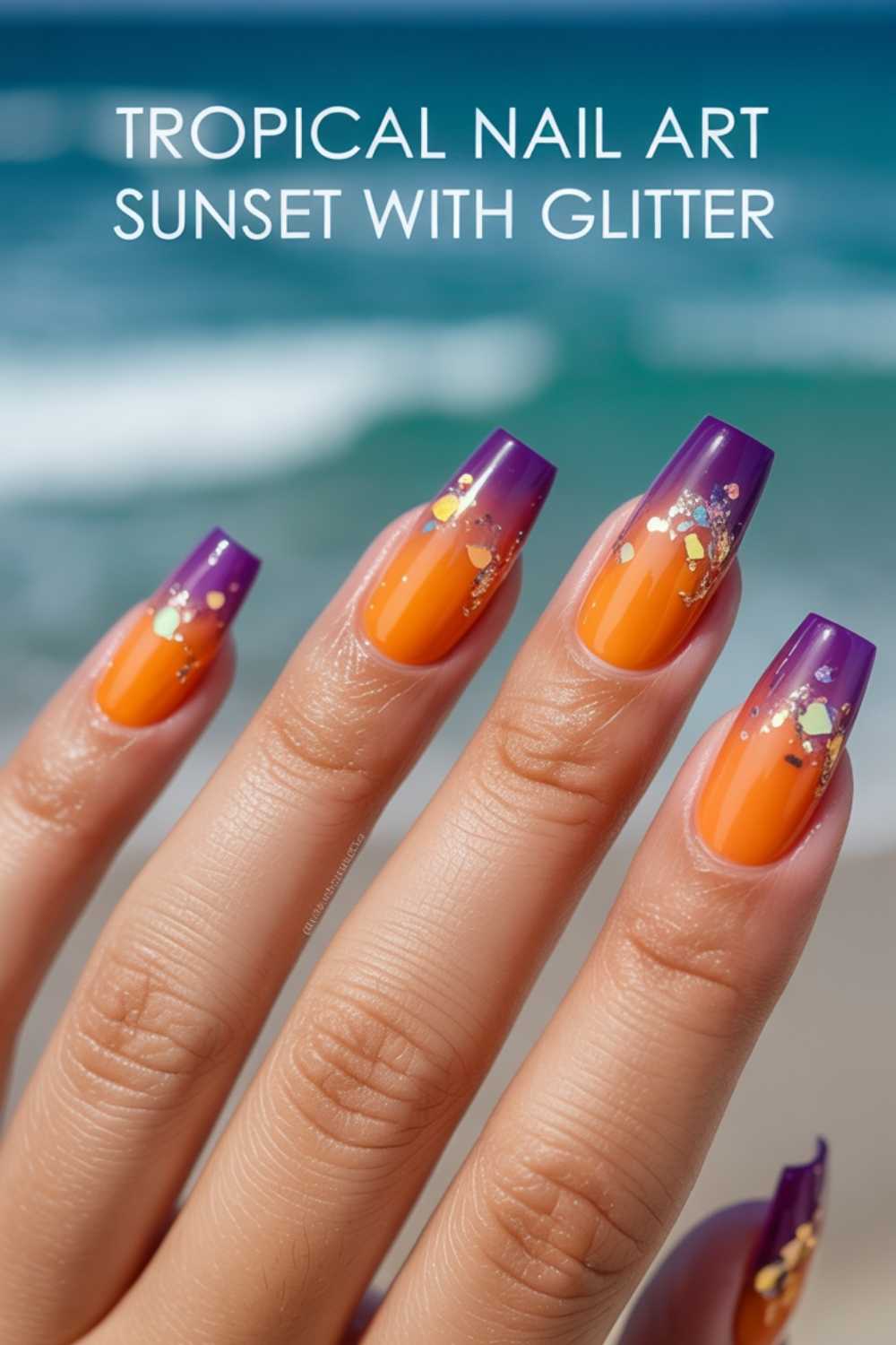 A hand with long nails painted in a tropical sunset nail art style features orange polish, purple tips, and gold glitter accents, set against a blurred beach background. Text reads: Tropical Nail Art Sunset With Glitter.