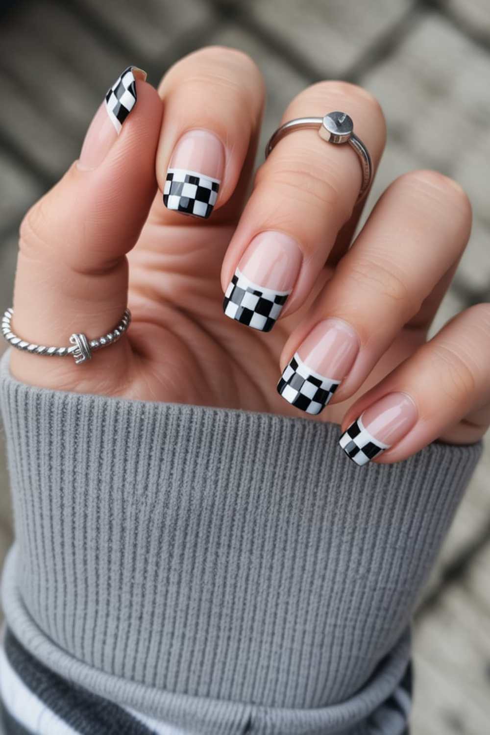 A hand with square-shaped nails featuring black and white checkerboard French tips. The person wears silver rings and a gray ribbed sleeve covers part of the hand. The background is slightly blurred.