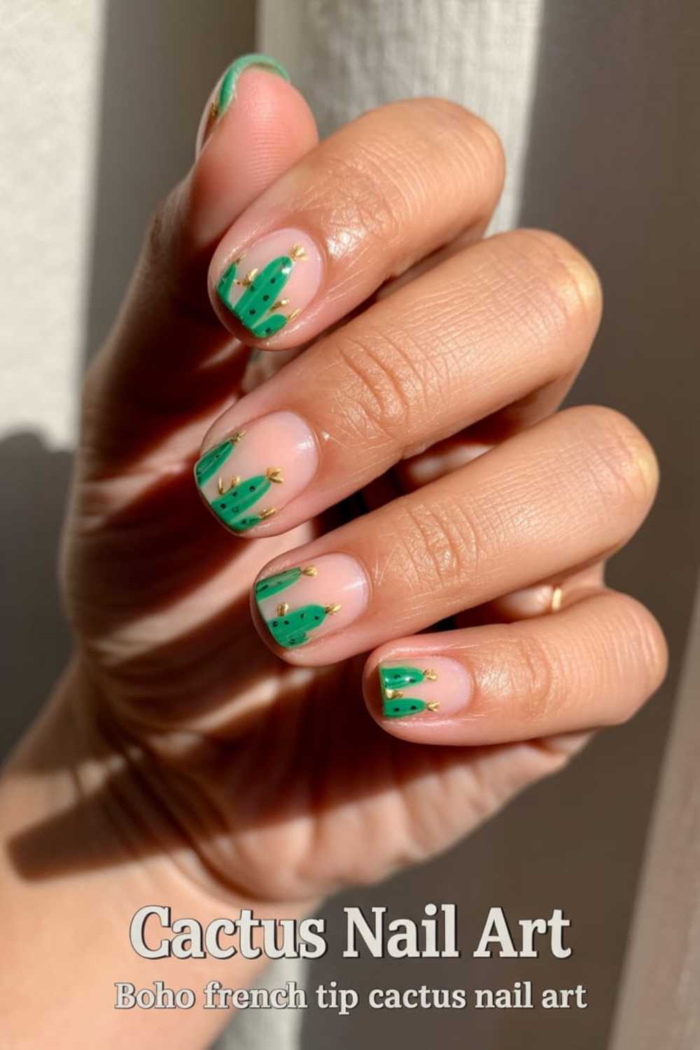 A hand with light brown skin shows nails painted with green cactus designs and small gold accents on a nude base; sunlight highlights the nails. Text on image reads, Cactus Nail Art: Boho french tip cactus nail art.