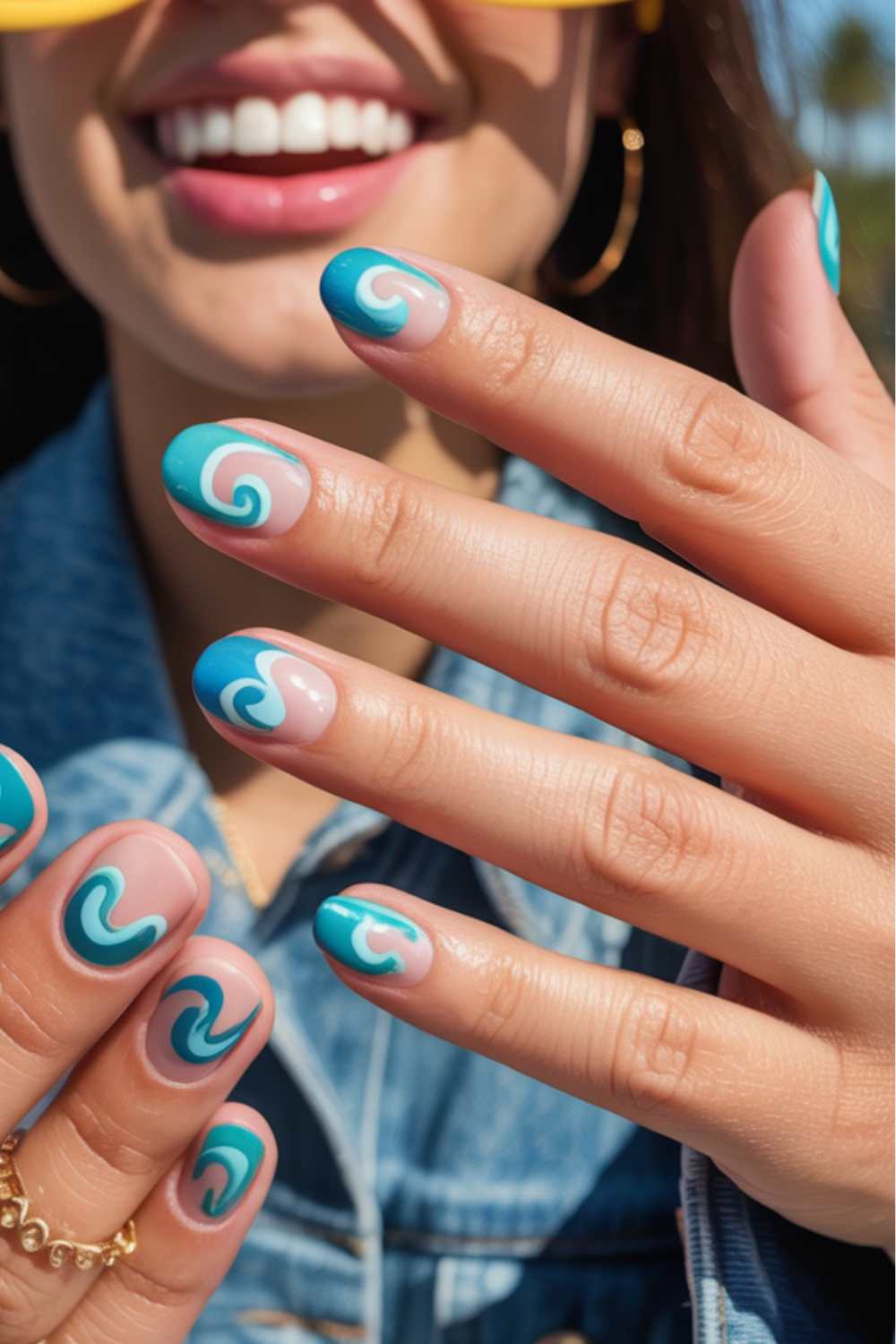 A smiling woman wearing gold hoop earrings and a denim jacket shows off her hands with teal and light pink swirl nail art designs. Her nails are medium-length and glossy.
