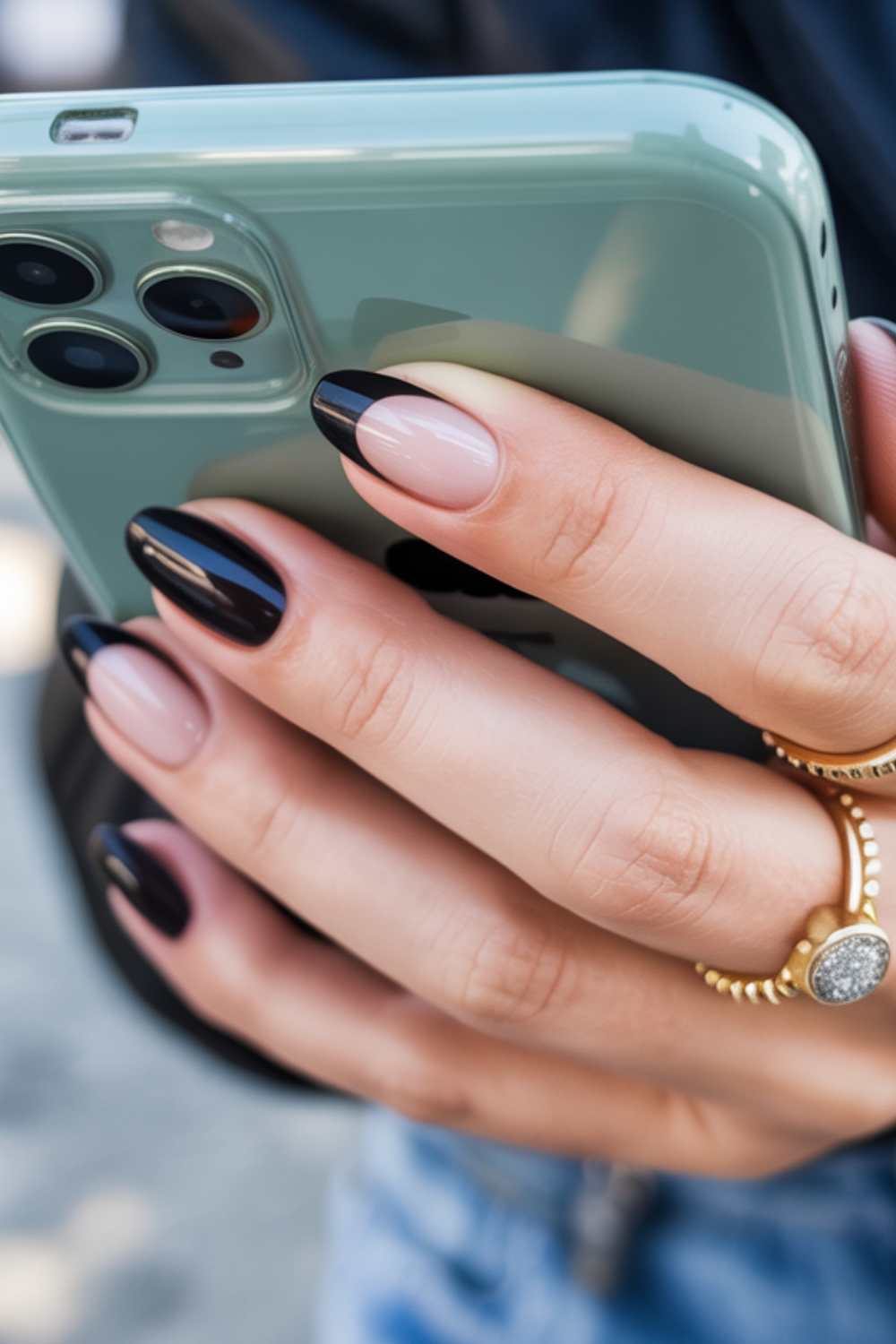 A close-up of a hand holding a green smartphone, showing nails painted with a stylish black and nude French tip design. The person is wearing gold rings, including one with a round, sparkly stone.